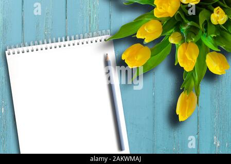 top view of blank spiral notepad, pencil and bouquet of yellow tulips on rustic blue wooden table template Stock Photo