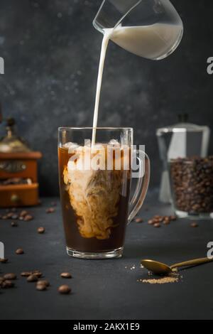 Iced latte coffee in cup glass with pouring milk on black. Close up. Stock Photo