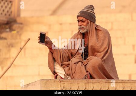 VARANASI, INDIA, JANUARY 18, 2019 : A man sitting along a ghat and facing the Ganges river, is holding a smartphone. Stock Photo