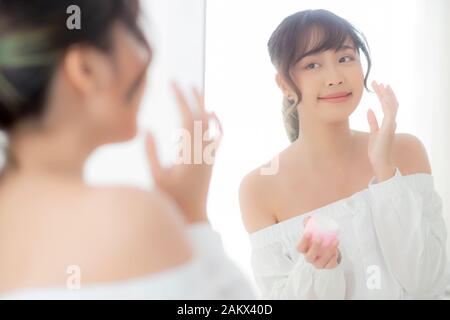 Beautiful portrait young asian woman applying cream moisturizer or lotion skin care cosmetic on face looking mirror, girl with treatment facial, healt Stock Photo