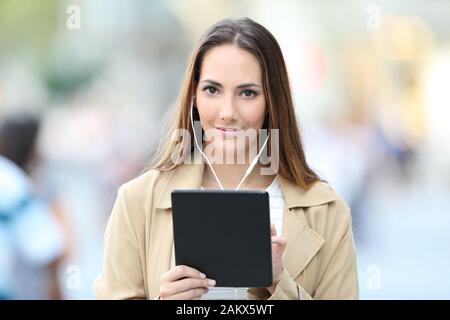 Fronf view portrait of a serious girl wearing earbuds holding a tablet looking at you in the street Stock Photo