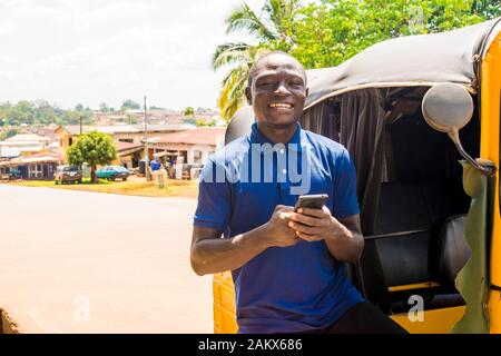 cheerful african man standing next to his tuk tuk taxi smiling and using his smart phone Stock Photo