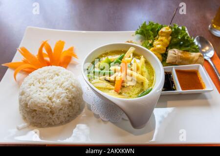 A bowl of traditional Thai green chicken curry with boiled rice, a satay, a spring roll and dips served in a china bowl on a white plate Stock Photo