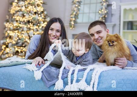 Young family of three and their lap-dog on sofa with Christmas background behind. Little boy playing with bed cover. Stock Photo