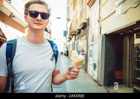 Caucasian young male tourist in dark sun glasses holding big italian ice cream in cone and walking on an european old town street during holidays. Tra Stock Photo