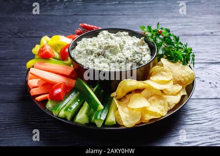 close-up of Creamy spinach dip in a bowl with sausages, carrots, cucumber, sweet pepper sticks, potato chips and fresh parsley on a plate