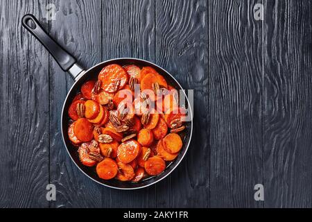 Candied sweet potatoes with brown sugar, maple syrup, orange juice and pecan nuts in a skillet on a black wooden table, horizontal view from above, fl Stock Photo