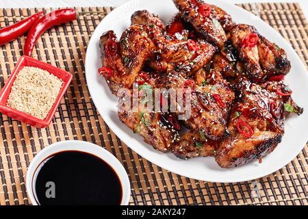 Oven-Baked Teriyaki Chicken Wings  sprinkled with sesame seeds, chili pepper pieces and parsley on a white plate on a bamboo mat, view from above Stock Photo