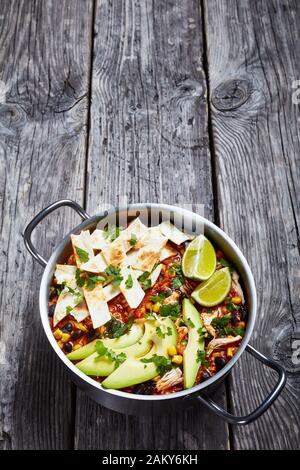 close-up of spicy Chicken Taco Soup with black bean, corn kernels, topped with tortilla strips, avocado slices and lime in a metal pot on a rustic woo Stock Photo