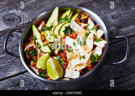 close-up of spicy Chicken Taco Soup with black bean, corn kernels, topped with tortilla strips, avocado slices and lime in a metal pot on a rustic woo Stock Photo