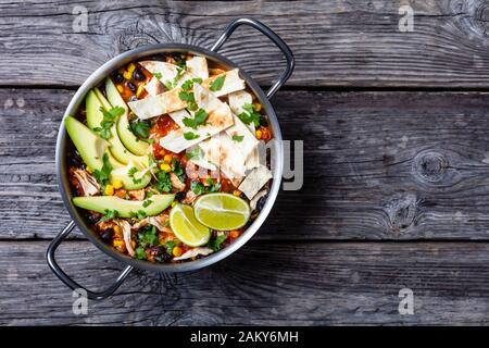 Chicken Taco Soup with black bean, corn kernels, topped with tortilla strips, avocado slices and lime in a metal pot on a rustic wooden table, flat la Stock Photo