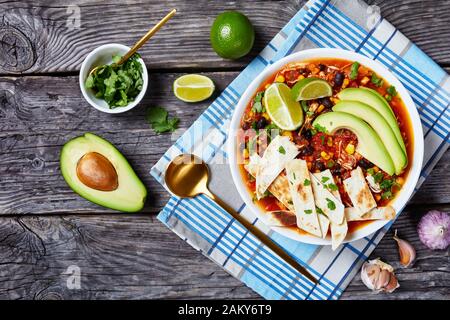 Chicken Taco Soup with black bean, corn kernels, topped with tortilla strips, avocado slices and lime in white bowl on a rustic wooden table, horizont Stock Photo