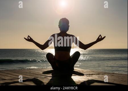 woman sitting by the sea practicing yoga and meditating, back view. Stock Photo