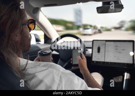 Young adult man sitting in car, using modern smartphone, drinking coffee Stock Photo