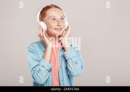 Inclusive Beauty. Girl with freckles in denim with headphones jacket standing isolated on grey listening music looking aside inspired copy space Stock Photo