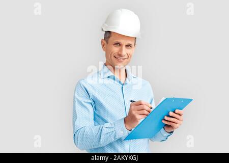 Construction. Mature man in hardhat standing isolated on white with clipboard taking notes smiling joyful Stock Photo