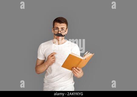 Freestyle. Young student in eyeglasses standing isolated on grey with book and party props mustache smiling confused Stock Photo