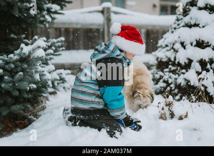 Boy in Santa hat playing in the snow with a dog in a yard. Stock Photo