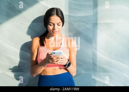 Young sporty woman sitting on mat, holding a phone in her hands, texting a message, having a break after practicing yoga, wearing black sportswear at Stock Photo