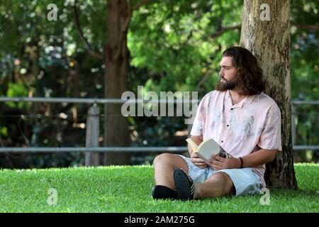 A young man is thinking about his studies, while he sits on the green grass at the park reading a book. Stock Photo