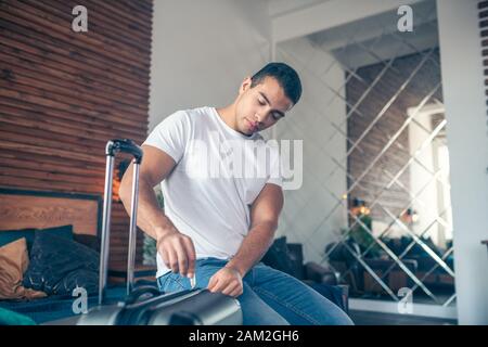 Dark-haired young man in white tshirt zipping his suitcase Stock Photo