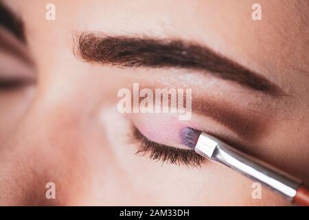 The work of a professional makeup artist. Eyeshadow applying, makeup for eyes closeup. Young woman applies pink colored eyeshadow with make up brush. Stock Photo