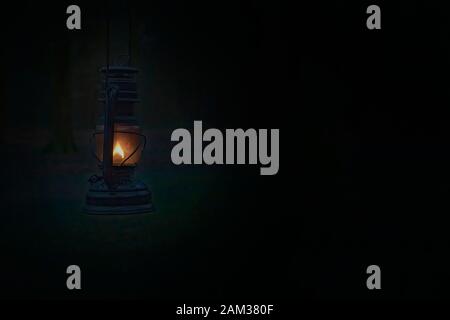 Vintage kerosene oil lantern lamp burning with a soft glow light in an dark forest / wood. Light in the darkness. Blurred background image of a glowin Stock Photo