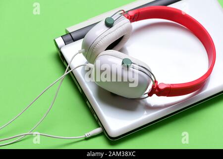 Headphones and silver laptop. Music and digital equipment concept. Electronics isolated on green background. Earphones in red and white colors made of plastic with computer. Sound recording idea. Stock Photo