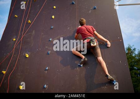 Sporty man practicing rock climbing in gym on artificial rock training wall outdoors. Young talanted slim climber guy on workout. Stock Photo