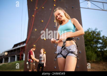 Pretty caucasian woman puts on belaying harness for practice on artificial rock wall outdoors. Slim strong healthy blonde prepares to conquer climbing Stock Photo