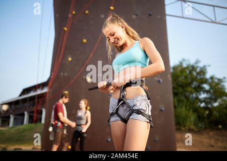 Pretty caucasian woman puts on belaying harness for practice on artificial rock wall outdoors. Slim strong healthy blonde prepares to conquer climbing Stock Photo