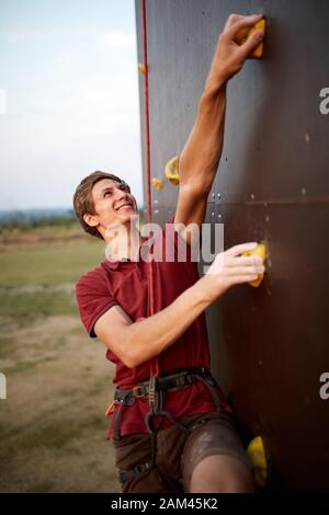 Sporty man practicing rock climbing in gym on artificial rock training wall outdoors. Young talanted smiling climber guy on workout. Stock Photo
