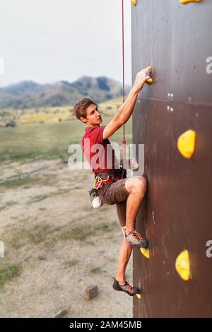 Sporty man practicing rock climbing in gym on artificial rock training wall outdoors. Young talanted climber guy on workout. Stock Photo