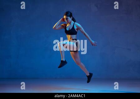 Caucasian young female athlete practicing on blue studio background in neon light. Close up of sportive model jumping high, running. Body building, healthy lifestyle, beauty and action concept. Stock Photo