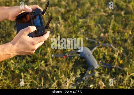 Man holds a drone remote controller in his hands. Quadcopter is on the ground ready to go and fly blurred on the background. Pilot is going to start Stock Photo