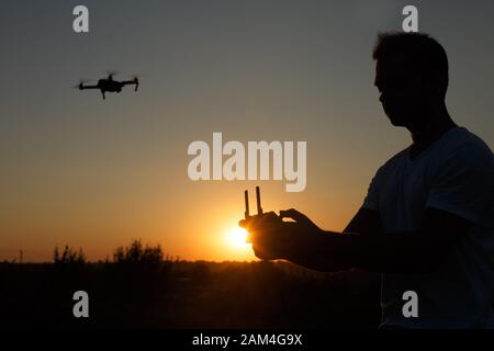 Silhouette of a man piloting drone in the air with a remote controller in his hands on sunset. Pilot takes aerial photos and videos with quadcopter Stock Photo