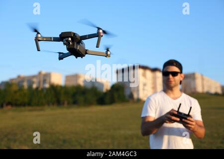 Compact drone hovers in front of man with remote controller in his hands. Quadcopter flies near pilot. Guy taking aerial photos and videos from above Stock Photo