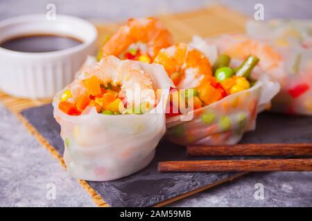 Spring rolls with vegetables and shrimps in a rice paper on the black plate. Near soy dip and chopsticks. Stock Photo