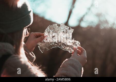 Back view of a blonde girl with a beanie holding a piece of thin ice towards the sun, admiring nature. Start of winter, cold temperatures. Stock Photo