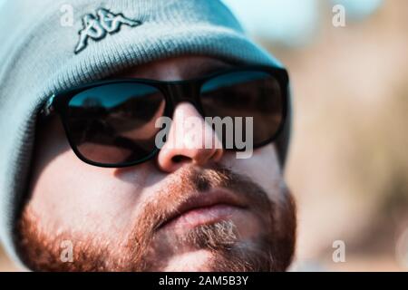 Closeup of a mans face, wearing sunglasses and a grey kappa logo beanie cleary visible. Orange beard Stock Photo