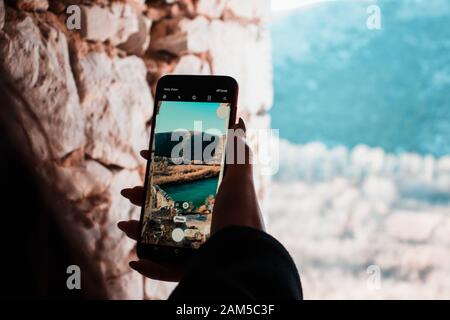 Woman's hand holding a smartphone taking a photo, looking out of a window of an old ancient building. River and scenery visible on the screen of the p Stock Photo