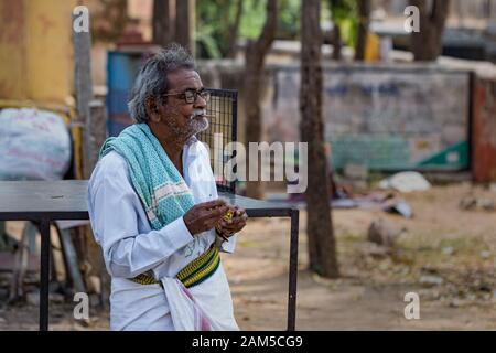 Hampi, Karnataka, INDIA - JANUARY 16, 2018: Elderly Indian man in glasses lights a cigarette. Resident of india. A man in a white shirt and dhoti Stock Photo