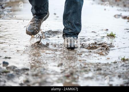 Man in sturdy brown walking boots splashing through a deep muddy mud puddle on a path after heavy rain in winter Stock Photo