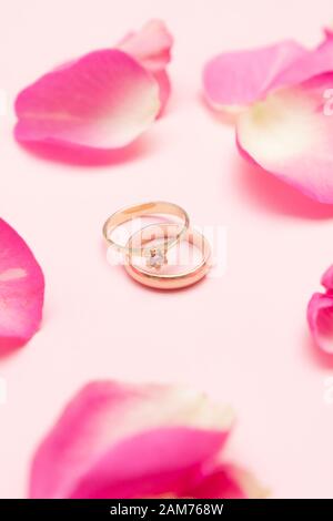 Wedding ring and gold engagement ring with gemstone around roses petals on the pastel pink background. Marriage concept. Happy Valentines day.