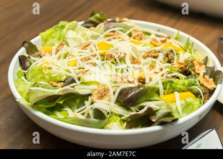 Closeup of delicious salad of lettuce, watercress, arugula, mango, walnuts and shredded cheese on white ceramic platter on wooden table. Healthy and n Stock Photo