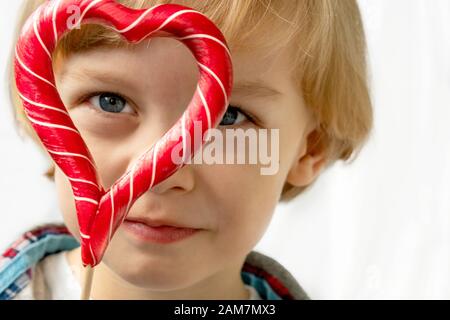 Cute little boy in with candy red lollipops in heart shape, white background. Beautiful kid eat sweets. Valentine's day, love concept. Stock Photo
