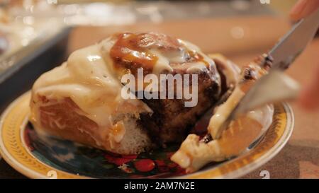 woman or man's hands cuts a fresh hot cinnamon bun spreaded with a white creamy-caramel cream on the beautiful plate in slow motion 4K video. Stock Photo