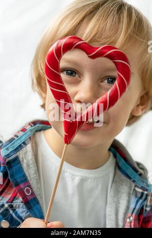 Cute little boy in with candy red lollipops in heart shape, white background. Beautiful kid eat sweets. Valentine's day, love concept. Stock Photo