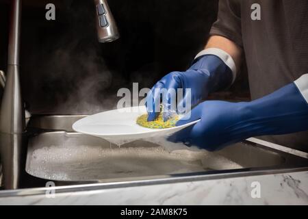 A model's hands washing dishes in steaming hot water with dark blue rubber gloves with anit-drip cuffs. Stock Photo