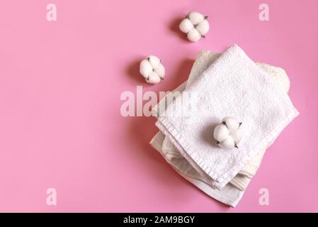 Stack of white cotton clothes and cotton flowers on pink background, flat lay, copy space. Organic cotton towels and clothes, eco lifestyle concept. Stock Photo